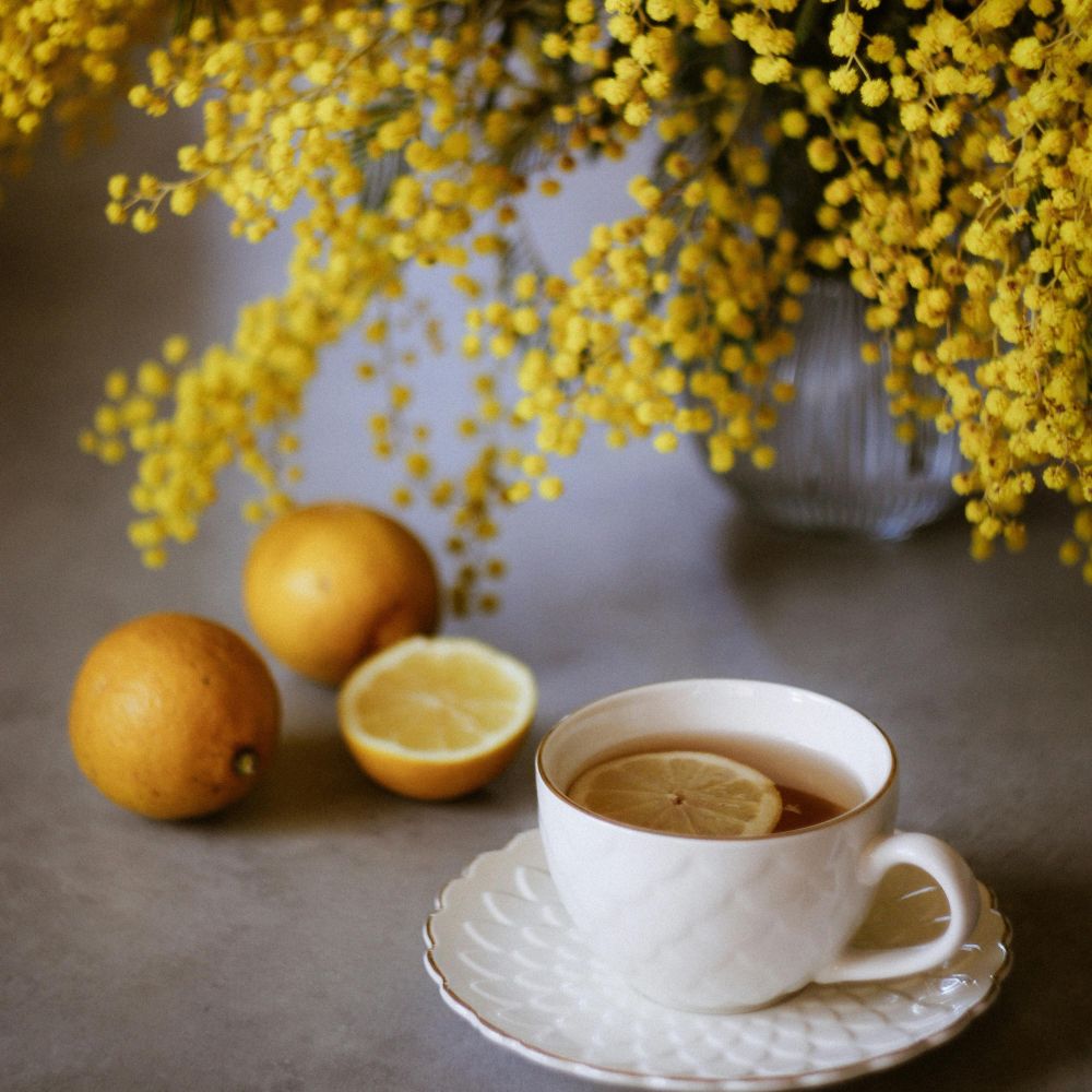 Ændringer i smags- og lugtesans symboliseret ved foto af hvid tekop med te og en skive citron. Ved siden af ligger et par citroner og en overskåret. Bag dette står en buket gule blomster med ser nederste del af.