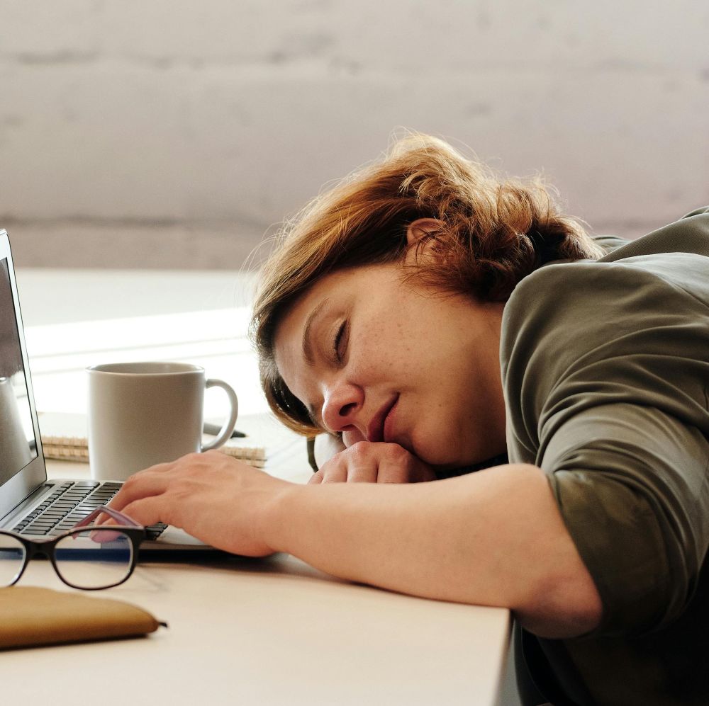 Kvinde faldet i søvn ind over sit arbejdsbord. Hun sidder ved en pc og med en kop kaffe stående ved siden af tastaturet. Brillerne ligger ved siden af.