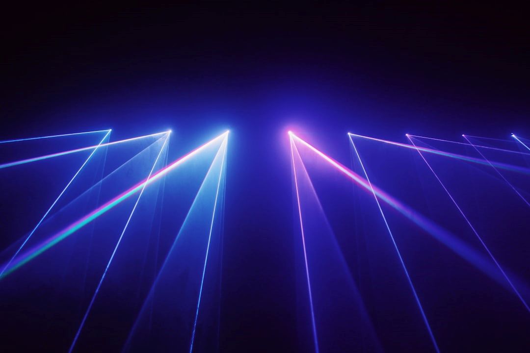 K-laser symboliseret ved lyskilder, der højt oppefra sender hvide og rosa laserstråler ned mod jorden i hver 3 stråler. Sort baggrund.