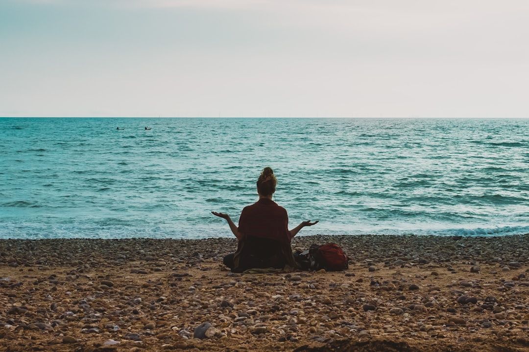 Meditation symboliseret ved kvinde der sidder i skrædderstilling på en strand foran et endeløst hav. Hun har armene vinklet ud til siden som hun modtager energi fra alt omkring hende.