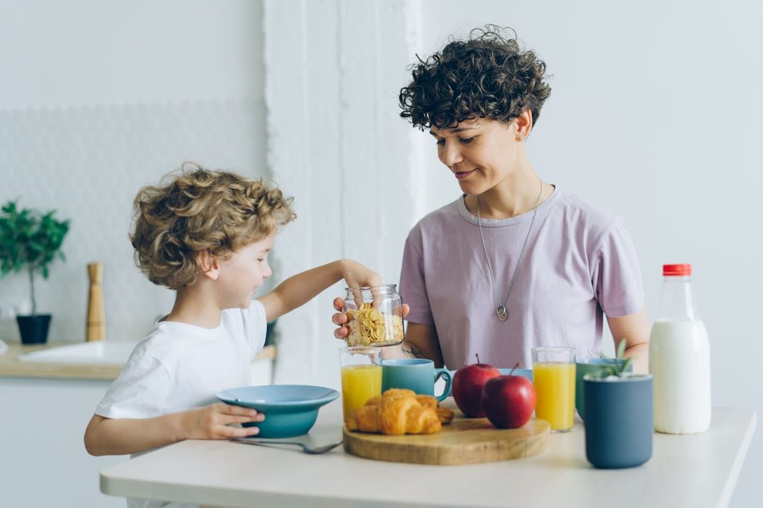 Kræftramte med mindreårige børn symboliseret ved ung mor, der står ved køkkenbord med barn på 6-7 år. Foran dem er arrangeret morgenmad med tallerkener, mælk, æbler, juice og croissant.