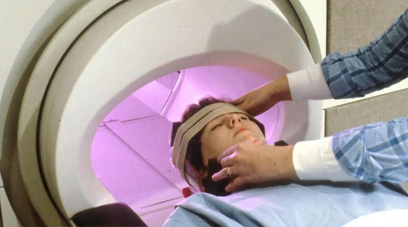 MR-scanning illustreret ved patient der ligger på ryggen på vej ind i en MR-scanner. Patienten får rettet lejring af hoved med to hænder fra person man kun ser underarme af.
