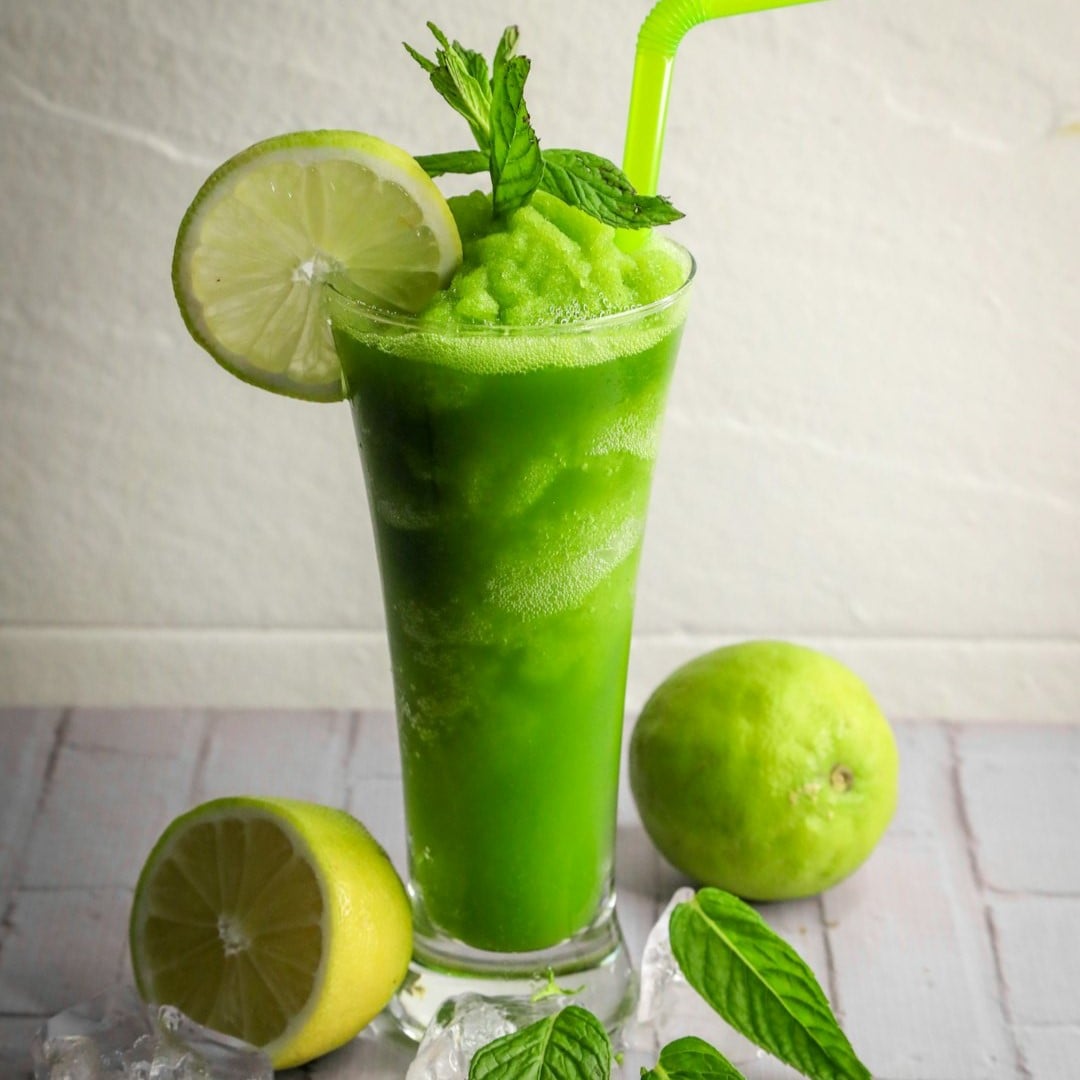 Højt glas med grønt indhold af grøntsagssmoothie. En skive citron over kanten på glasset og ½ citron ligger foran.