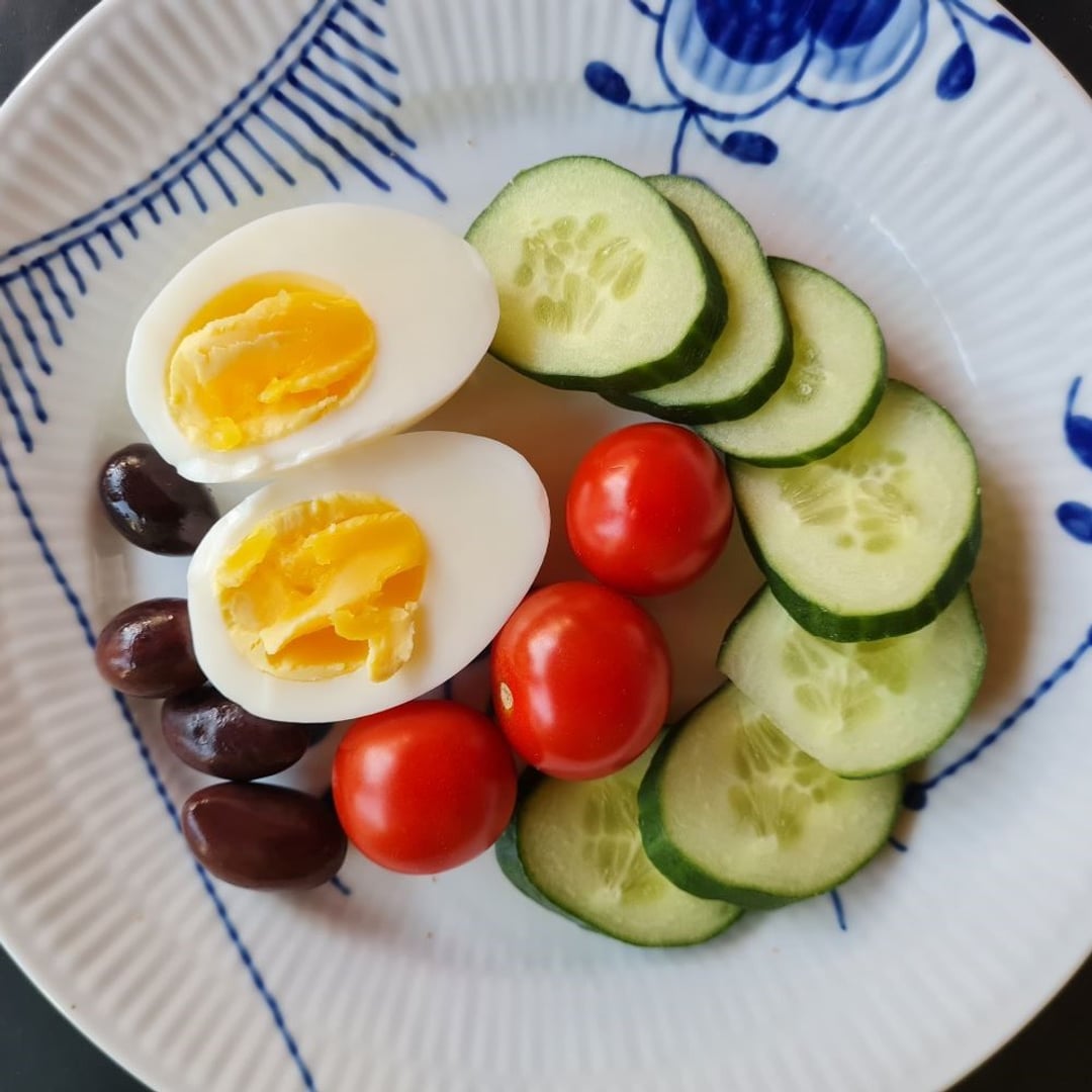 Et kogt æg - halveret, en halvkrans af agurkeskiver, 3 tomater i midten og 4-5 kalamata-oliven ligger op ad ægget.