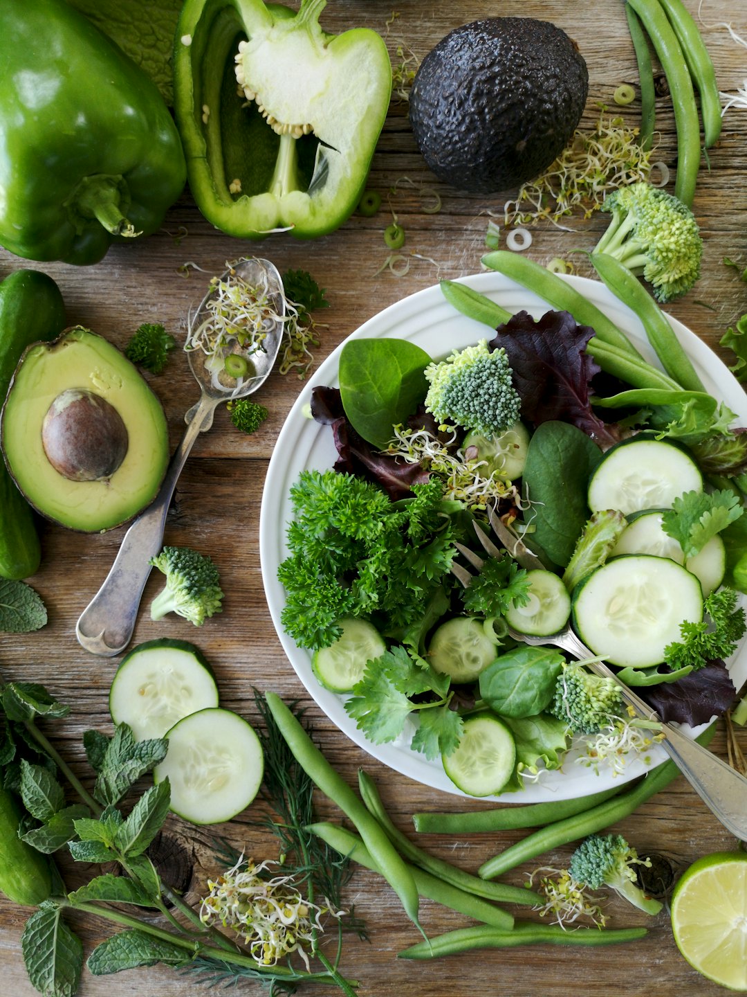 Raw Food symboliseret ved en tallerken med skiver af rå agurk, squash, grønne bønner og forskellig slags salat og friske spirer. Ved siden af ligger halve avocado, overskåret grøn peber, agurkeskiver og en gammel enkel sølvske.