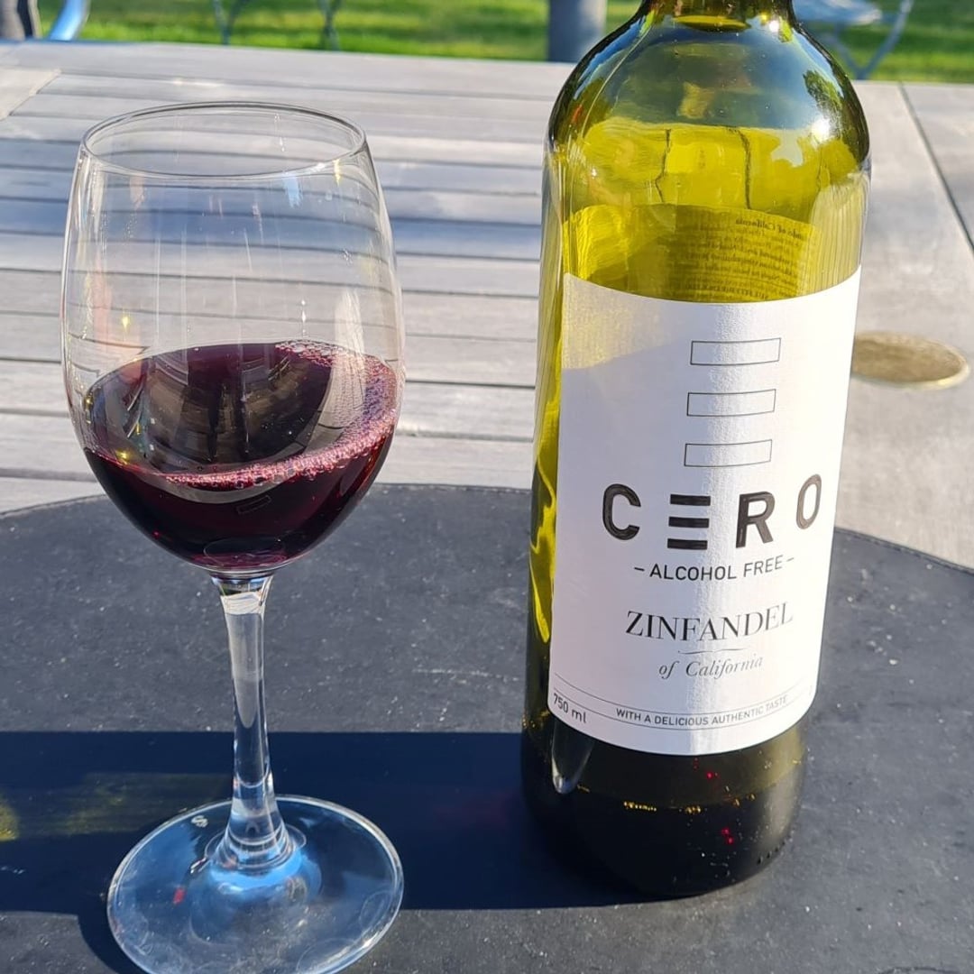 Foto af glas med rødvin på terassebord, man ser lille del af. Ved siden af glasset står en flaske med påskriften Cero - Zinfandel.