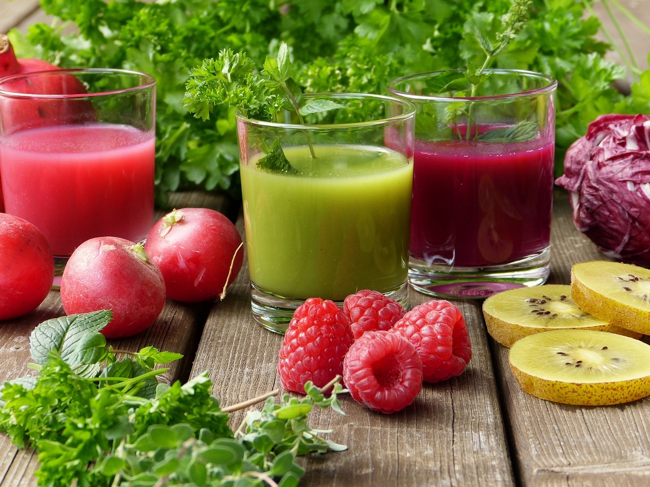 Detox symboliseret ved træbord, hvorpå der står et glas med rød grøntsagsjuice, et glas med grøn grøntsagsjuice og et med mørkerød juice. Ved siden af ligger salat, skiver af kiwi, hindbær, granatæbler og krydderurter.