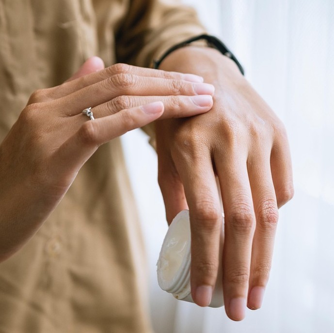 Personlig pleje og kosmetik symboliseret ved hænder, hvor den ene holder en krukket med creme og den anden smører creme på overfladen af hånden.