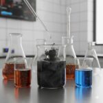 Aktivt kul symboliseret ved nogle bægerglas og reagensglas der står på et bord. De indeholder væska med blå og brune farver. I midten står et bægerglas med kul.