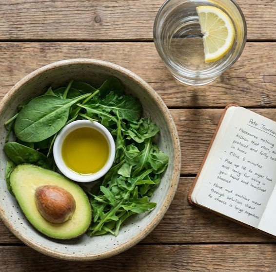 Pete Sulaks Protokol symboliseret ved en skål med bladgrønt og ½ avocado samt olie, et glas vand med citron og en notesbog med optegnelser.