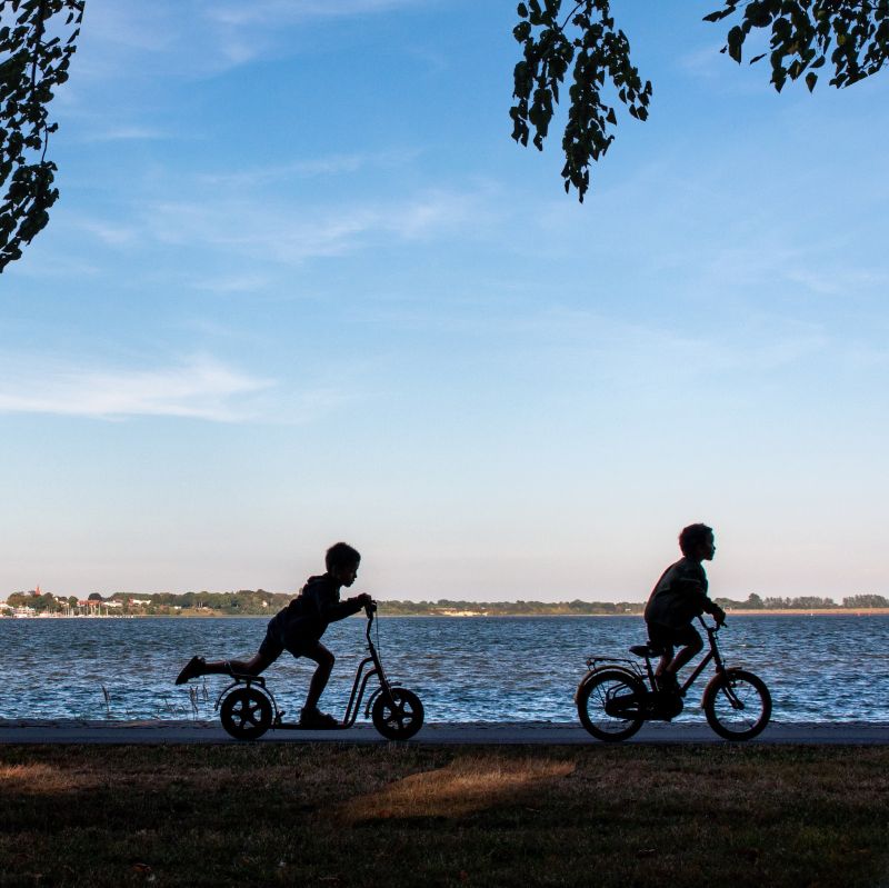 Super Seje Søskende Dag symboliseret ved silhuet af to børn der cykler. Vand i baggrunden.